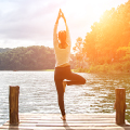 Person stands in tree pose on a dock by a lake, surrounded by forest in bright sunlight.